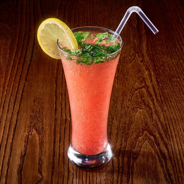A tall glass of pink Indian drink with mint, lemon, and a straw on a wooden table.